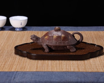 May include: A brown ceramic teapot shaped like a turtle, with a lid and a spout. The teapot is sitting on a wooden base with a brown and black design. The teapot is on a brown woven mat.