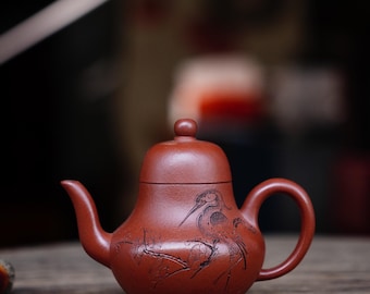 May include: A reddish-brown ceramic teapot with a bird and branch design. The teapot has a curved spout, a rounded handle, and a small lid with a knob. The design is etched into the surface of the teapot.