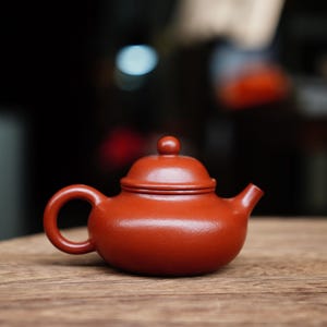 May include: A small, reddish-brown ceramic teapot with a rounded body, a curved handle, and a small spout. The lid has a small knob on top. The teapot sits on a wooden surface, with a blurred background.