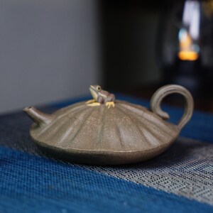 Puede incluir: Una tetera de cerámica marrón con una figurita de rana en la tapa. La tetera tiene un cuerpo redondeado con una superficie texturizada y un pico curvo. La tetera está sentada sobre una superficie con un patrón azul y blanco.