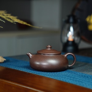 Peut inclure: Une théière en céramique brun foncé avec un couvercle et une poignée. La théière est posée sur une surface à motifs bleus et verts.
