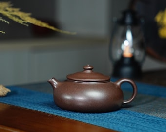 May include: A dark brown ceramic teapot with a lid and a handle. The teapot is sitting on a blue and green patterned surface.