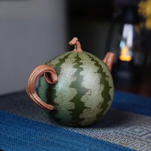 May include: A green and brown ceramic teapot shaped like a watermelon. The teapot has a handle and spout.