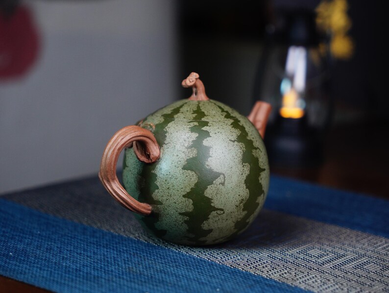 May include: A green and brown ceramic teapot shaped like a watermelon. The teapot has a handle and spout.