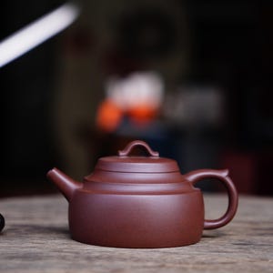 May include: A small, reddish-brown ceramic teapot with a curved handle and a lid. The teapot is on a wooden surface. The spout is small and angled. The background is blurred, with a dark, out-of-focus appearance.