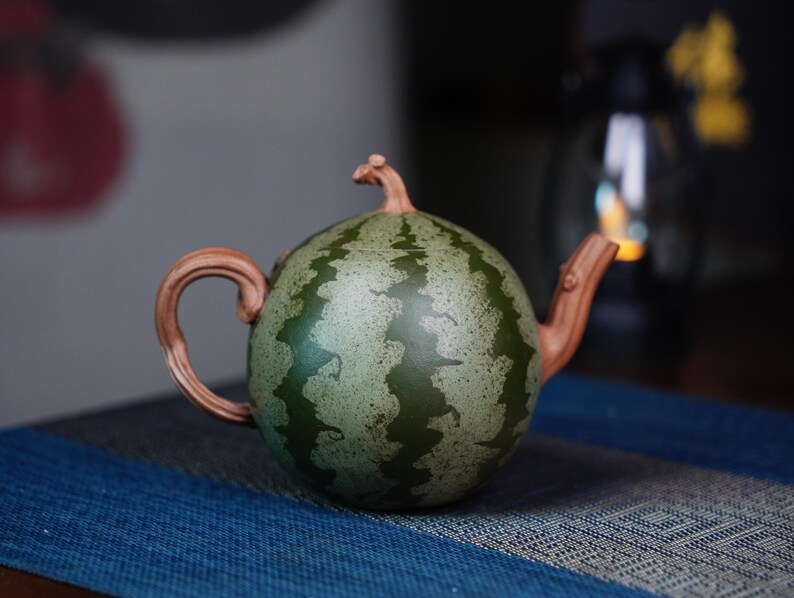 May include: A green and brown ceramic teapot shaped like a watermelon. The teapot has a handle and a spout.
