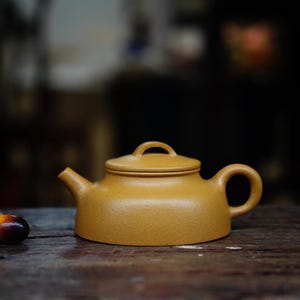 May include: A yellow ceramic teapot with a rounded body and a lid. The teapot has a small handle on the side and a spout on the front.
