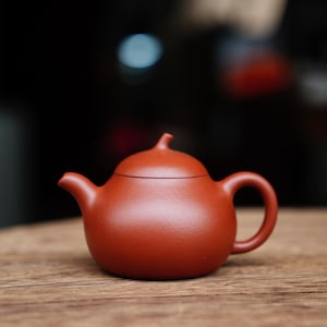 May include: A small, reddish-brown ceramic teapot with a rounded body, a curved spout, and a loop handle. The lid has a small knob. The teapot sits on a wooden surface, with a dark background.