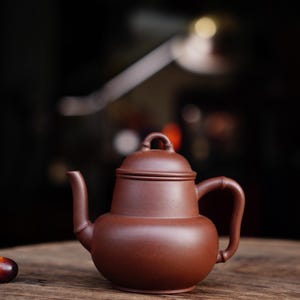 以下が含まれることがあります： 蓋と取っ手のある茶色の陶器製のティーポット。ティーポットは木製のテーブルの上に置かれています。