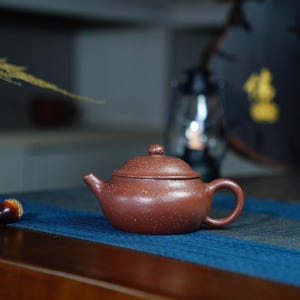 Op de afbeelding: Een klein, bruin, keramisch theepotje met een deksel en een handvat. Het theepotje heeft een gespikkelde textuur en staat op een blauw oppervlak.