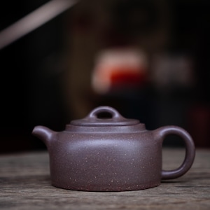 May include: A small, brown ceramic teapot with a rounded body, a curved spout, and a loop handle. The lid has a small, round knob. The teapot is sitting on a wooden surface.