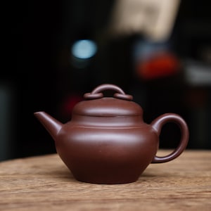 Könnte beinhalten: Kleine, braune Keramik-Teekanne mit rundem Körper, gebogenem Ausguss und Schlaufengriff. Der Deckel hat einen kleinen, gewölbten Griff. Die Teekanne steht auf einer Holzoberfläche mit verschwommenem Hintergrund.