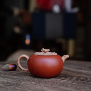 May include: A small, round, reddish-brown teapot with a matching lid and handle. The lid has a carved wooden accent. An acorn rests on the wooden surface next to the teapot. The background is blurred.