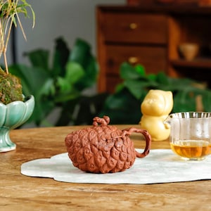 Puede incluir: Naturaleza muerta con una tetera marrón texturizada con tapa a juego, una jarra de vidrio pequeña con té y una pequeña figura amarilla. También hay una planta verde en un cuenco decorativo. Los objetos están sobre una mesa de madera.