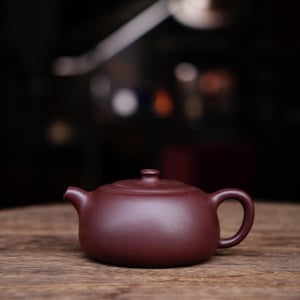 May include: A dark red clay teapot with a rounded body and a small spout. The teapot is sitting on a wooden table.