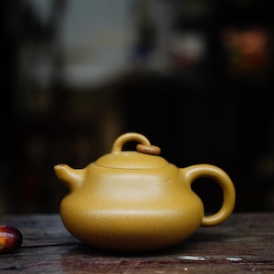 May include: A mustard-yellow ceramic teapot with a rounded body, curved handle, and a small spout. The lid has a wooden accent. The teapot sits on a dark wooden surface, with a blurred background.