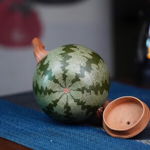 May include: A green and brown ceramic teapot with a watermelon pattern. The teapot has a wooden handle and a matching lid.