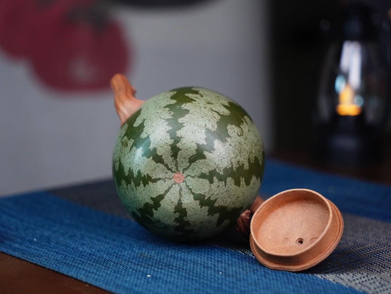May include: A green and brown ceramic teapot with a watermelon pattern. The teapot has a wooden handle and a matching lid.