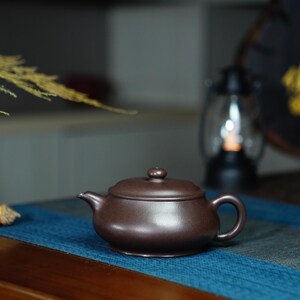 May include: A dark brown ceramic teapot with a lid and a handle. The teapot is sitting on a blue table runner.