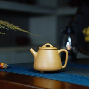 以下が含まれることがあります： 丸みを帯びたボディと小さな注ぎ口を持つ、薄い茶色のセラミック製のティーポット。ティーポットは青い表面の上に置かれています。