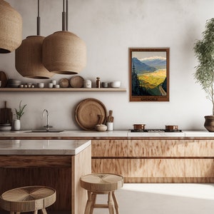 May include: A vintage travel poster for Grenoble, France, depicting a scenic mountain road winding through a valley with a town in the distance. The poster is framed and hanging on a wall in a modern kitchen with wooden cabinets and a white worktop.