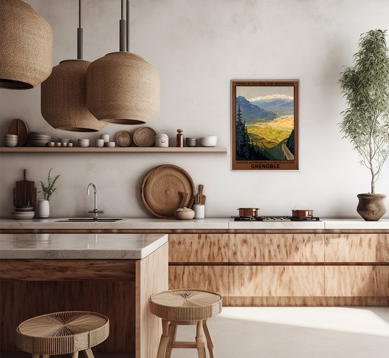 May include: A vintage travel poster for Grenoble, France, depicting a scenic mountain road winding through a valley with a town in the distance. The poster is framed and hanging on a wall in a modern kitchen with wooden cabinets and a white worktop.