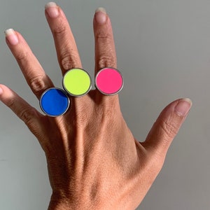 May include: Three colorful rings on a hand. The rings are round and have a silver band. The rings are blue, yellow, and pink.