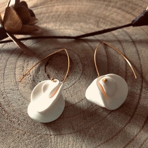 Pode incluir: Um par de brincos de flores de cerâmica branca com detalhes dourados pendurados em um fio dourado.