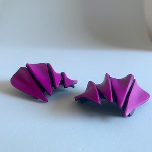Op de afbeelding: Een paar abstracte, geometrische oorbellen van paars plastic met zwarte accenten. De oorbellen zijn gevormd als een reeks overlappende driehoeken.