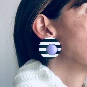 Pode incluir: Um par de brincos grandes e redondos com um design listrado preto e branco. O centro de cada brinco tem um círculo roxo.