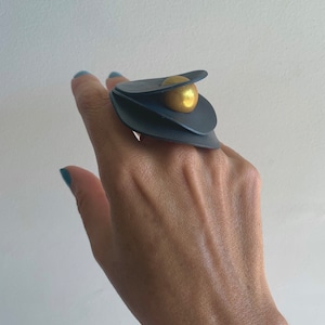 Pode incluir: Um anel preto com uma esfera dourada no centro. O anel é feito de várias camadas de material preto.