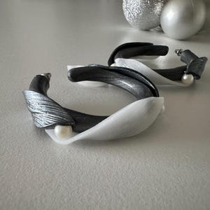 Pode incluir: Um par de brincos artesanais com um design único. Os brincos apresentam uma combinação de elementos prateados, pretos e brancos, com um detalhe de pérola. O design é abstrato e moderno, adequado para uma variedade de ocasiões.