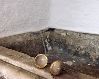 May include: A pair of stud earrings. Each earring has a light-coloured, rounded dome with a gold-coloured rim. The earrings are displayed on a textured, light-coloured surface, with a white wall in the background. The earrings are a jewelry item.