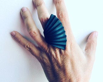 May include: A large, black, fan-shaped ring with a textured surface. The ring is worn on the index finger of a hand.