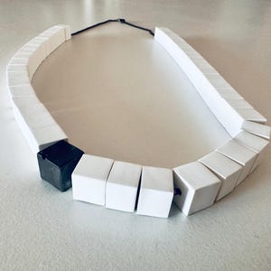 Pode incluir: Um colar moderno com uma série de contas brancas em forma de cubo conectadas por um cordão preto. Uma única conta preta em forma de cubo é incorporada no design. O colar é exibido em uma superfície clara.