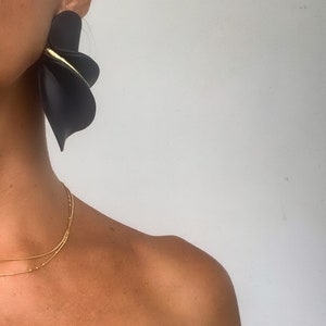 May include: A woman wearing a black off-the-shoulder top and a gold chain necklace. She is wearing large black statement earrings with a gold accent.