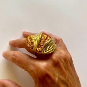 Pode incluir: Um anel dourado marcante com uma seção central texturizada e tecida, ladeada por dois elementos estilizados em forma de leque e canelados. O anel é usado em uma mão contra um fundo branco, mostrando seu design arrojado e acabamento metálico.