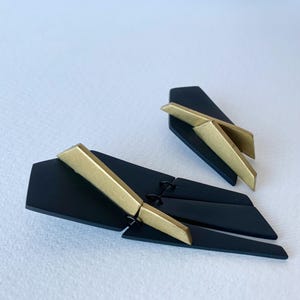 May include: A pair of geometric earrings in black and gold. The earrings are made of two pieces, one larger than the other, that are connected by a small metal hinge. The larger piece has a gold accent in the shape of a lightning bolt.