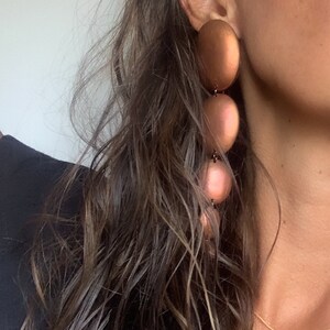May include: A woman wearing a black top with a gold chain necklace and large, round, copper-colored earrings.