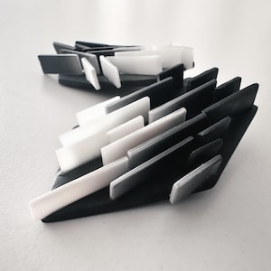 Può includere: Un paio di orecchini caratterizzati da una base angolare nera decorata con molteplici elementi rettangolari sporgenti in varie tonalità di bianco, grigio e nero, creando una struttura geometrica tridimensionale a strati con finitura opaca.
