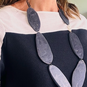 Pode incluir: Um colar marcante com grandes contas cinzentas em forma oval. As contas são conectadas por pequenas ligações metálicas. O colar é usado sobre um top preto e branco. Uma joia única.