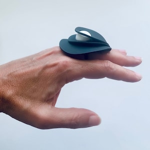 Könnte beinhalten: Ein schwarzer Ring mit einem weißen perlmuttartigen Zentrum. Der Ring besteht aus mehreren überlappenden Teilen, die ein einzigartiges und modernes Design schaffen.