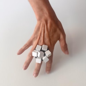 May include: A contemporary ring with a cluster of white square blocks on a black base. The ring has a striking geometric design. The ring is displayed on a hand against a white backdrop.