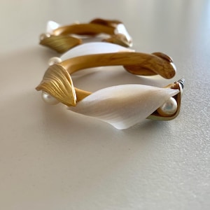 Pode incluir: Uma pulseira única com elementos dourados em forma de folha, detalhes brancos em forma de pétala e pequenas pérolas redondas. A pulseira tem uma aparência delicada e artística, adequada para ocasiões especiais.