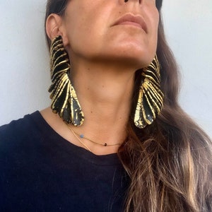 Peut inclure: Une paire de grandes boucles d'oreilles statement avec une base noire et des accents dorés. Les boucles d'oreilles sont composées de plusieurs couches de fines pièces superposées, créant un aspect texturé et dimensionnel.