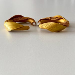 Pode incluir: Um par de brincos dourados com um design orgânico único. Os brincos apresentam uma seção texturizada e com contas e uma forma suave semelhante a uma pétala. Cada brinco tem um pequeno detalhe de pedra preciosa transparente.