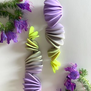 Könnte beinhalten: Ein Paar lange, farbenfrohe Ohrringe aus geschichtetem Polymerton. Die Ohrringe sind ein Farbverlauf von Lila, Grau und Gelb. Die Ohrringe werden vor einem weißen Hintergrund mit lila Blumen gezeigt.