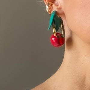 Pode incluir: Brincos de cereja. Cada brinco apresenta uma cereja vermelha brilhante com folhas verdes e uma haste dourada. Os brincos são usados na orelha de uma pessoa, com um brinco de argola dourada visível.