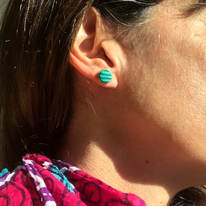 Pode incluir: Um close-up de um brinco de botão redondo com uma borda dourada. O brinco apresenta um design listrado turquesa e verde-azulado. O brinco é usado na orelha de uma pessoa com cabelo castanho.