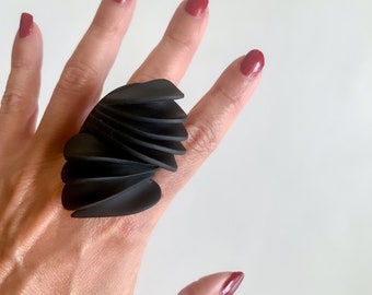 Puede incluir: Un anillo negro, abstracto, escultórico con un diseño orgánico en capas. El anillo se lleva en un dedo.